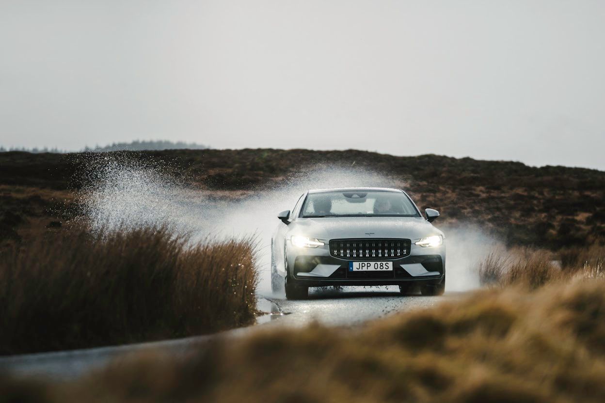A grey Polestar 1 driving on a wet road surrounded by brown grass.