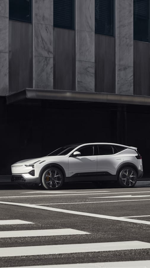 A white Polestar 3 SUV is parked on a street in front of a tall building with a dark facade.