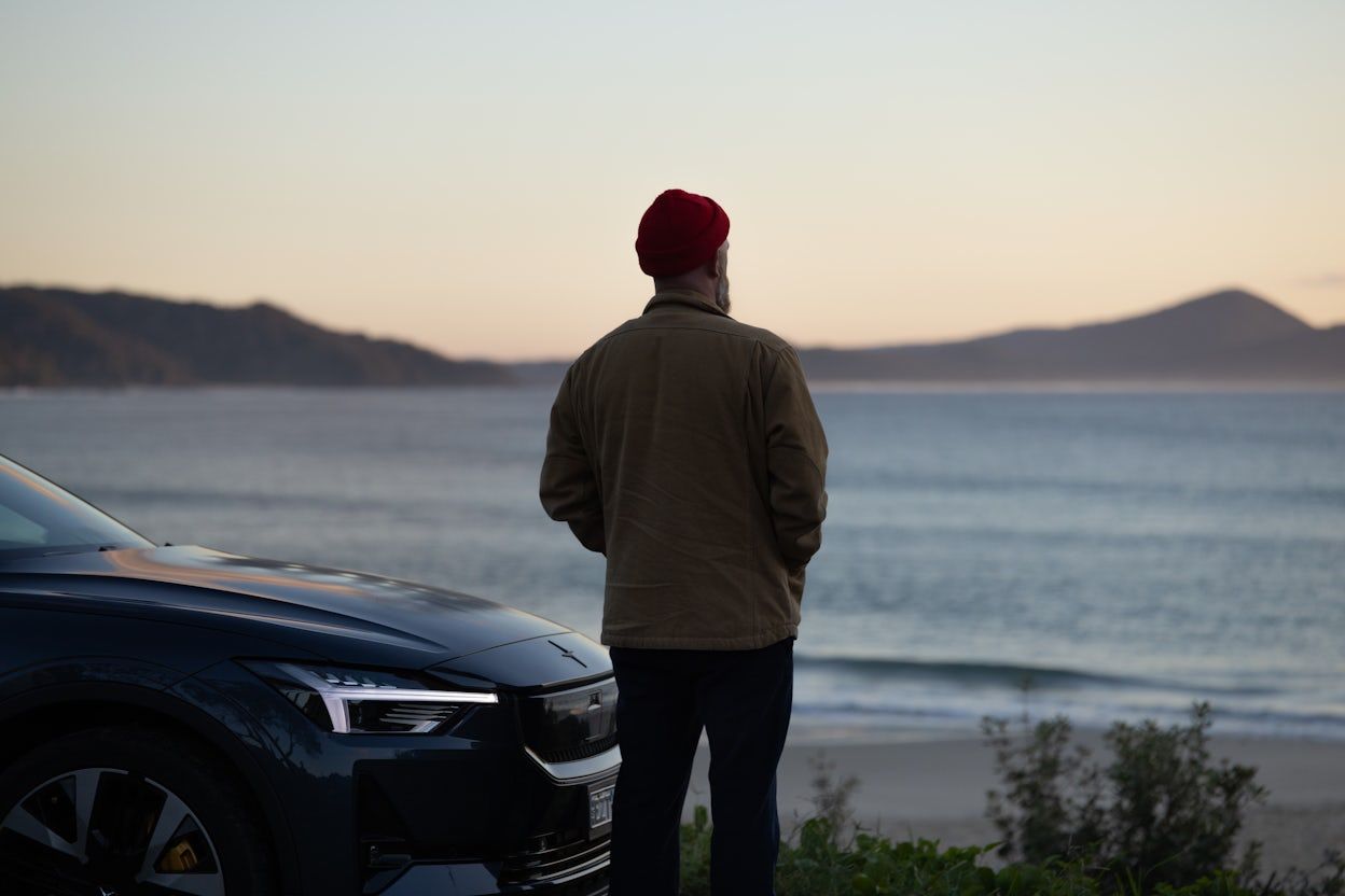 Man stares out to the ocean from the beach at dawn, next to the bonnet of a Polestar 2