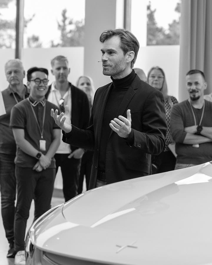 A man in a suit speaks to a group of people gathered around a white car inside a modern building.