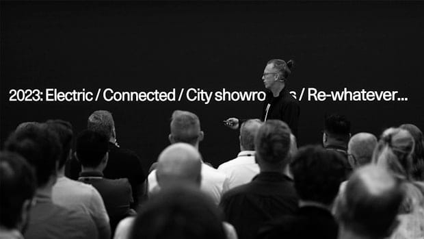 A person with a topknot hairstyle stands on a stage in front of a seated audience, presenting a slide with the text "2023: Electric / Connected / City showroom / Re-whatever...