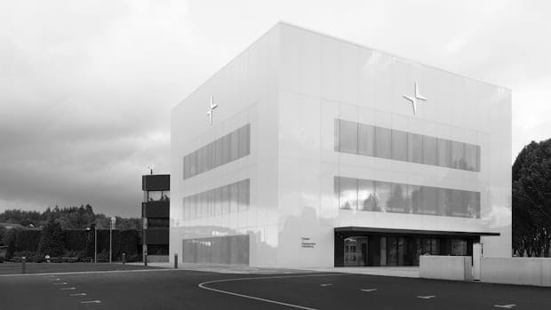 Polestar HQ, a modern, white cubic building with a minimalist design and Polestar logo on the upper corners, surrounded by a paved area and greenery under a cloudy sky.