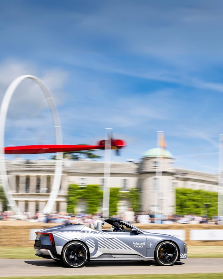 A Polestar 6 Concept driving, with the roof down past a large building, with a blurred view of a large statue and grass in the foreground.