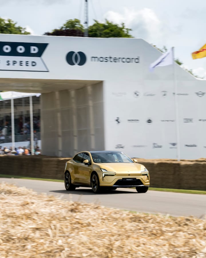 A Swedish gold Polestar 4 driving past a large building, with a blurred view of a large statue and grass in the foreground.