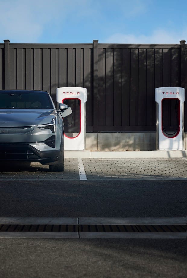 A Polestar 3 parked in front of Tesla charging stations against a dark wooden fence, under a clear blue sky.