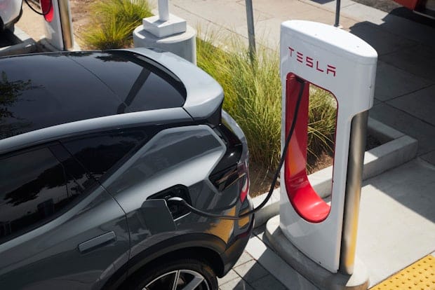 Polestar 3 car charging at a Tesla Supercharger station, surrounded by greenery and pavement.